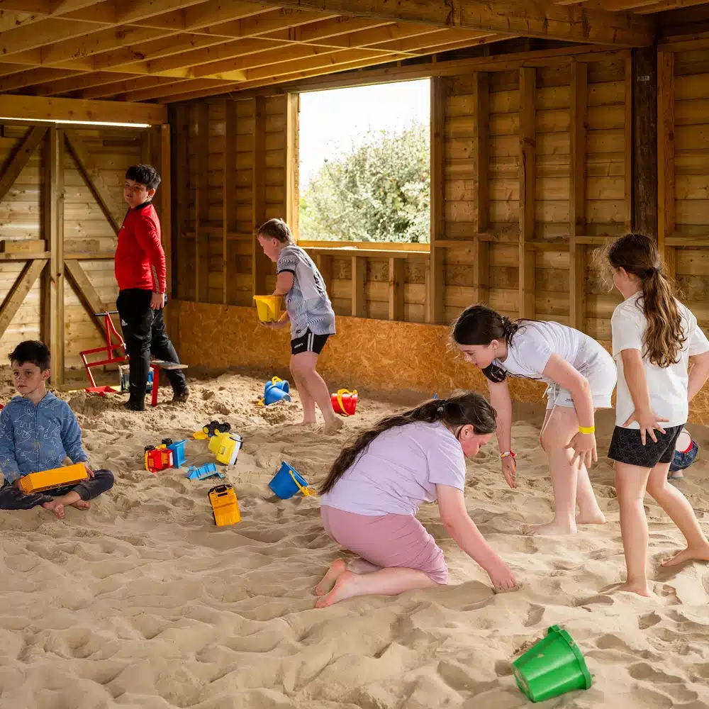 Six children play in an indoor sandy area, using colourful buckets and toy lorries. The wooden space, often used for Navan bouncy castle party fun, is filled with natural light as the children remain engaged and active.