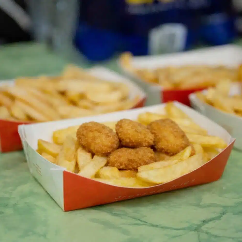 A tray of thick-cut chips topped with breaded chicken nuggets sits on a green table, ready to fuel your Navan roller disco party. In the background, two more trays promise plenty of deliciousness for every guest.