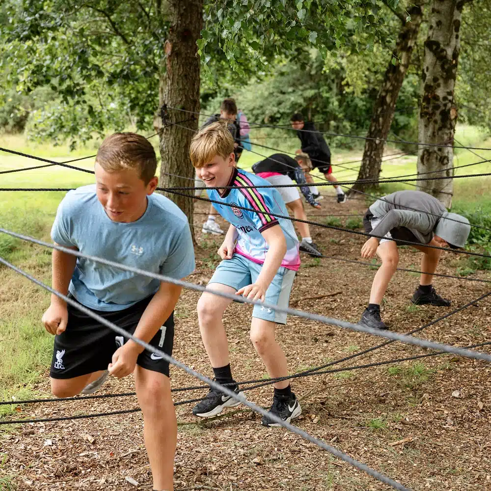 A group of boys are outdoors, weaving through a web of black ropes tied between trees during a Navan pick and mix party, participating in an obstacle course activity on a grassy, wooded area.