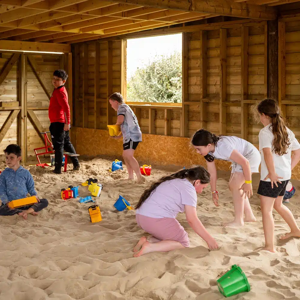 Children play in an indoor sand area with wooden walls at a Navan pick and mix party. Some kids dig and use buckets, whilst others sit or stand. Colourful toys and buckets are scattered as sunlight streams through the window and doorway.