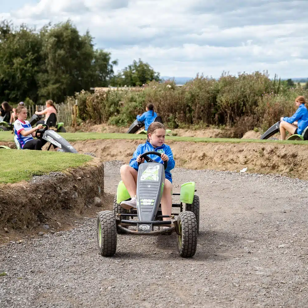 Children ride pedal-powered go-karts on an earth track outdoors, surrounded by grass and bushes. The sky is partly cloudy, and the children appear focused and engaged—perfect fun for a Navan pick 'n' mix party.