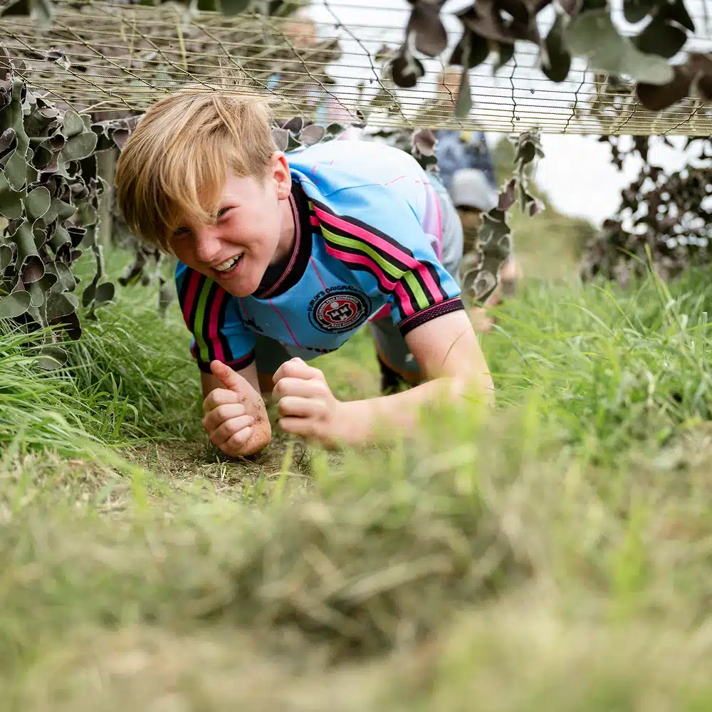 A smiling child in a striped sports shirt crawls through grass under a net at a Navan pick and mix party’s outdoor obstacle course, surrounded by greenery and leaves.