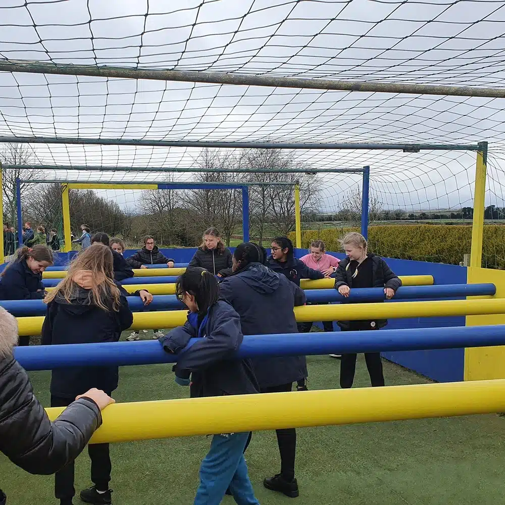 A group of children play human table football outdoors, holding onto colourful bars within a fenced, netted area at a Navan pick and mix party. Trees and hedges frame the fun under a cloudy sky.