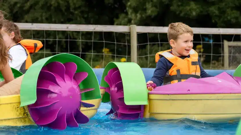 Two children wearing orange life jackets enjoy colourful pedal boats in a shallow pool at NeuroFest Navan. One child turns a pedal, smiling, as trees and a wooden fence create a cheerful backdrop.