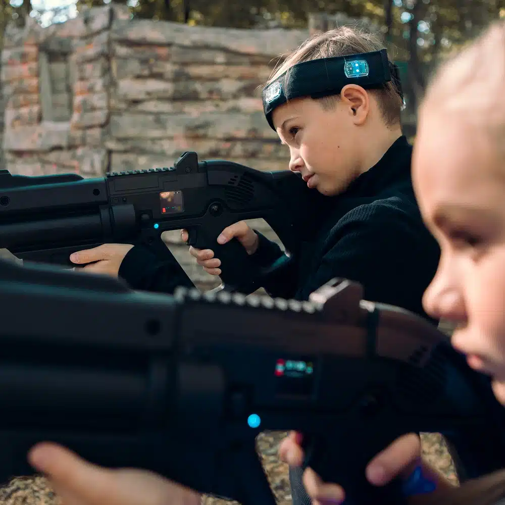 Two children wearing headbands and holding laser tag guns aim carefully whilst playing an outdoor laser tag game, with a wooden structure and trees in the background.