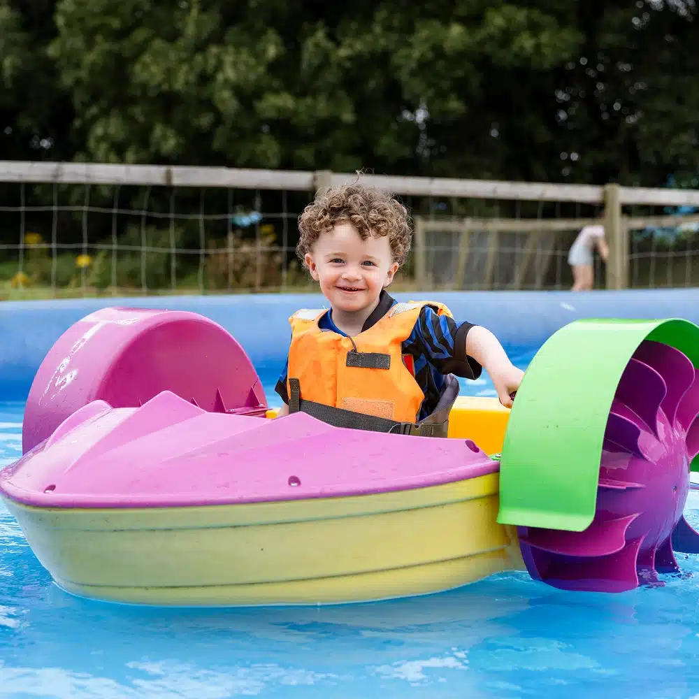 A young child wearing an orange life jacket smiles whilst sitting in a colourful paddle boat on a small pool, with trees and a fence in the background.