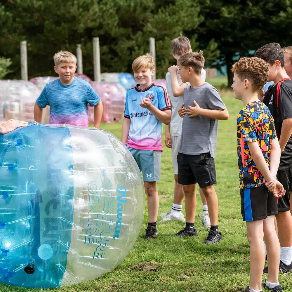 A group of boys stand outdoors on grass, smiling and talking near large inflatable bubble balls. One boy is partially inside a blue and clear bubble ball, while others watch and laugh. Trees and more bubbles are in the background.