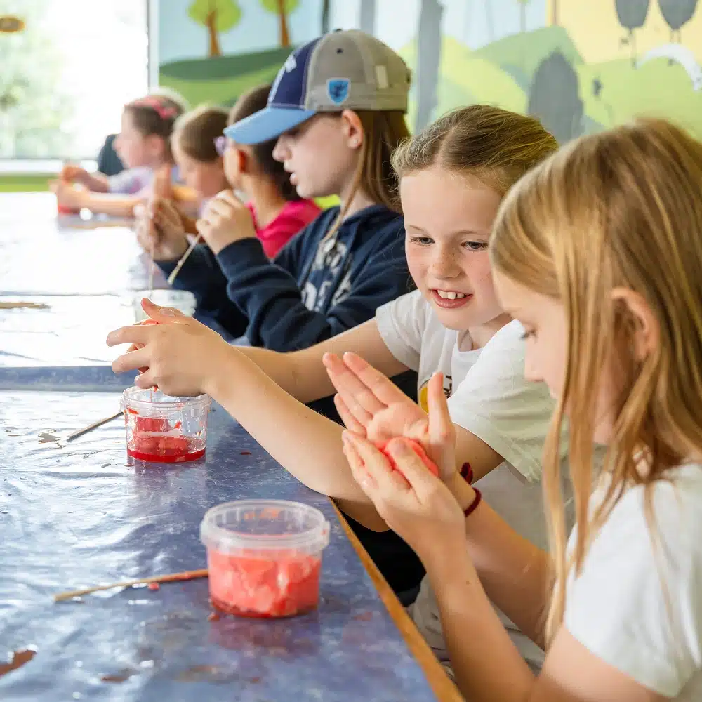 Children sit at a table playing with colourful slime, smiling and chatting. The table is covered with a blue cloth, and the background features a bright, mural-painted wall.