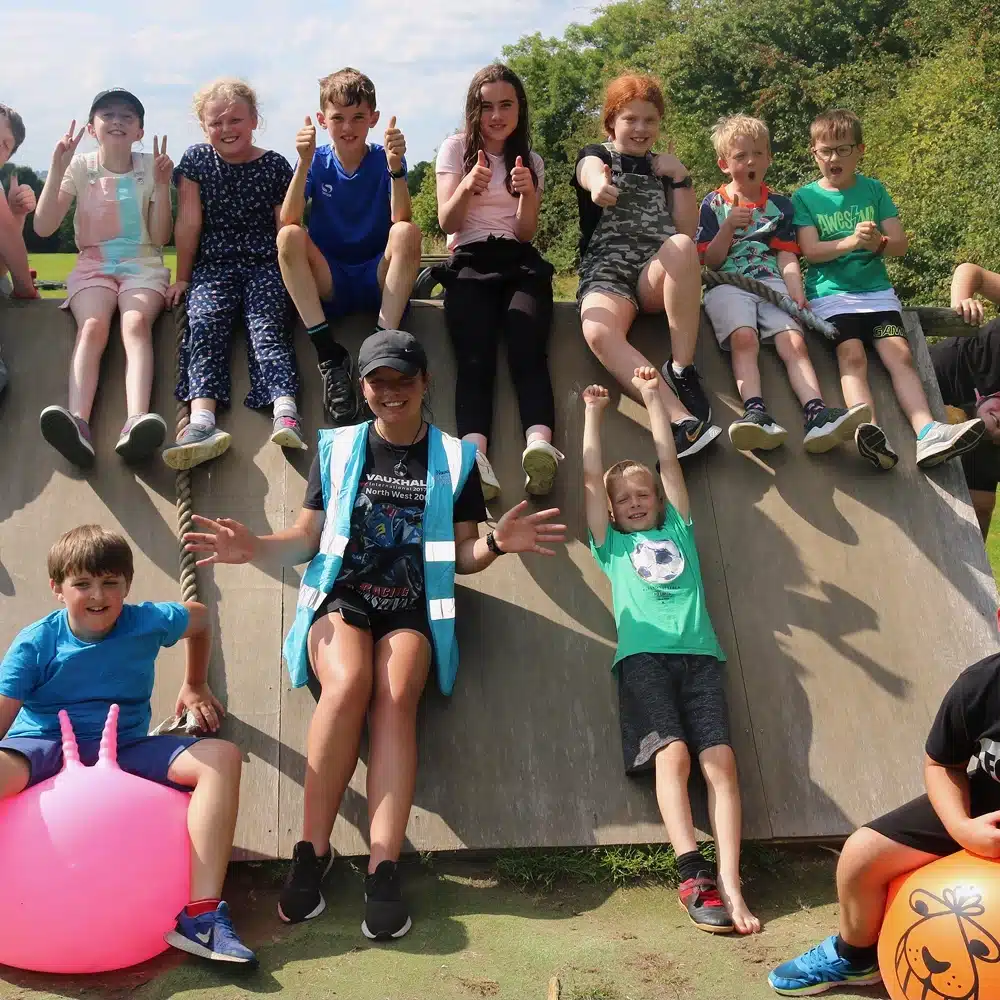 A group of children sit and play on a sloped wooden structure outdoors, smiling and posing for the camera on a sunny day. An adult in a blue waistcoat sits at the bottom, surrounded by kids.