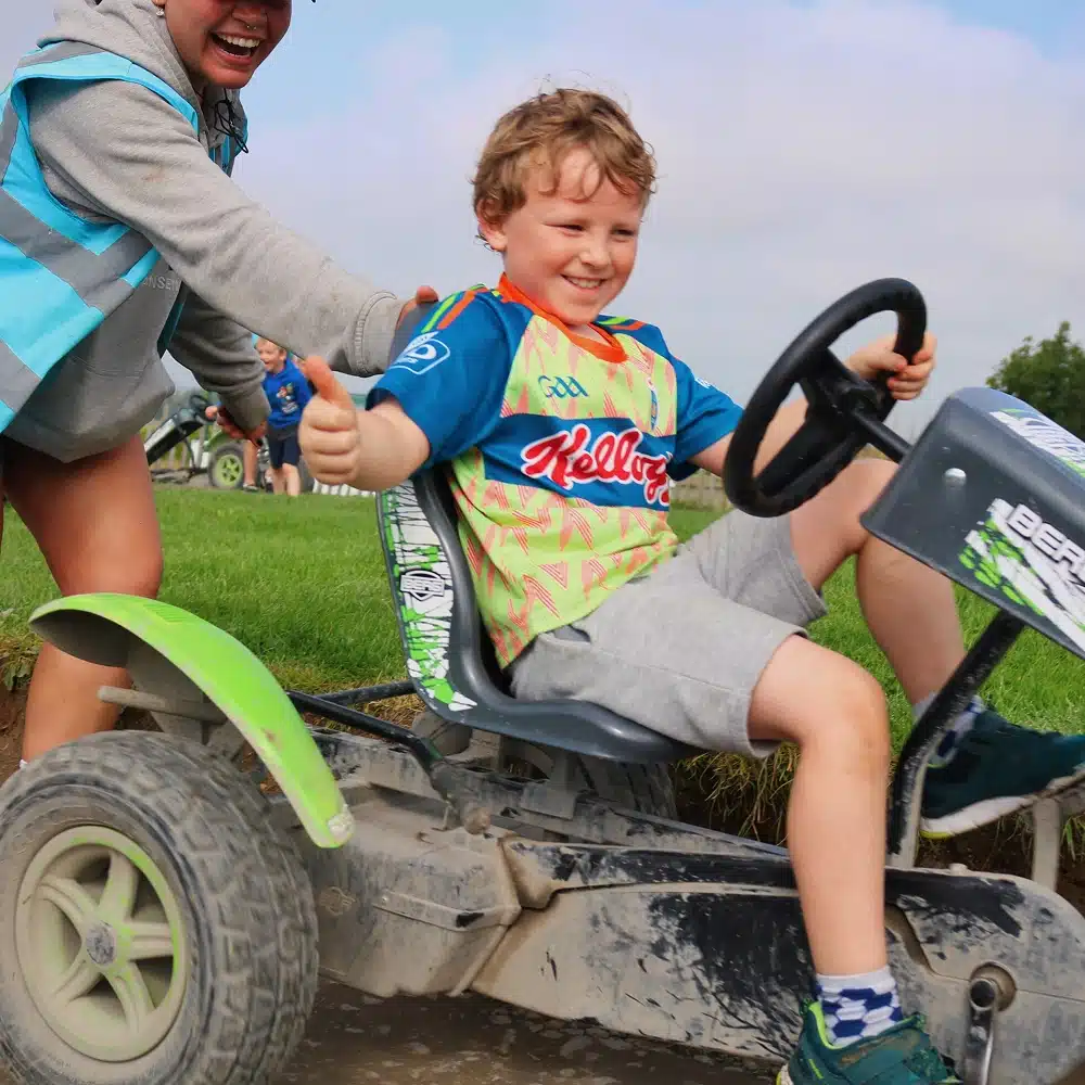 A smiling child drives a small go-kart on a dirt track while an adult pushes from behind, both appearing joyful and energetic. Grass and more people are visible in the background.