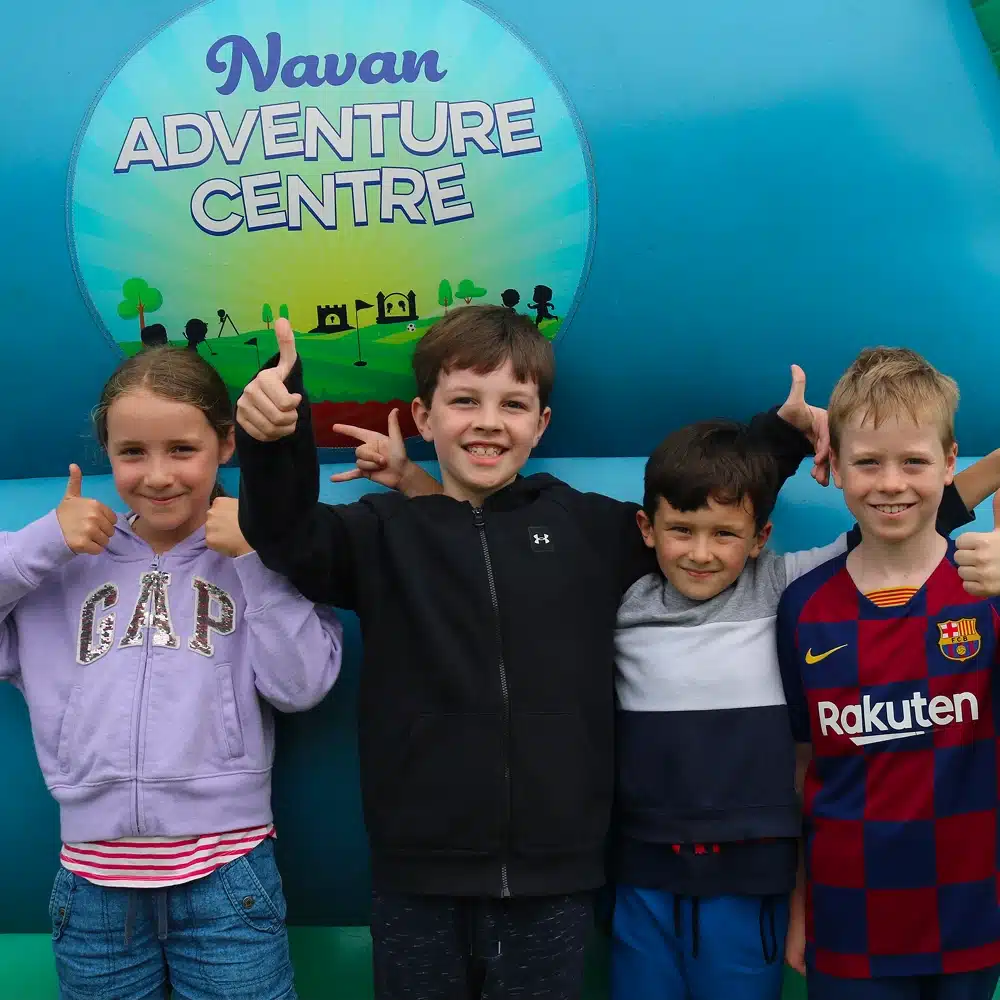 Four smiling children stand in front of a blue sign that reads "Navan Adventure Centre," giving thumbs up and posing happily together.