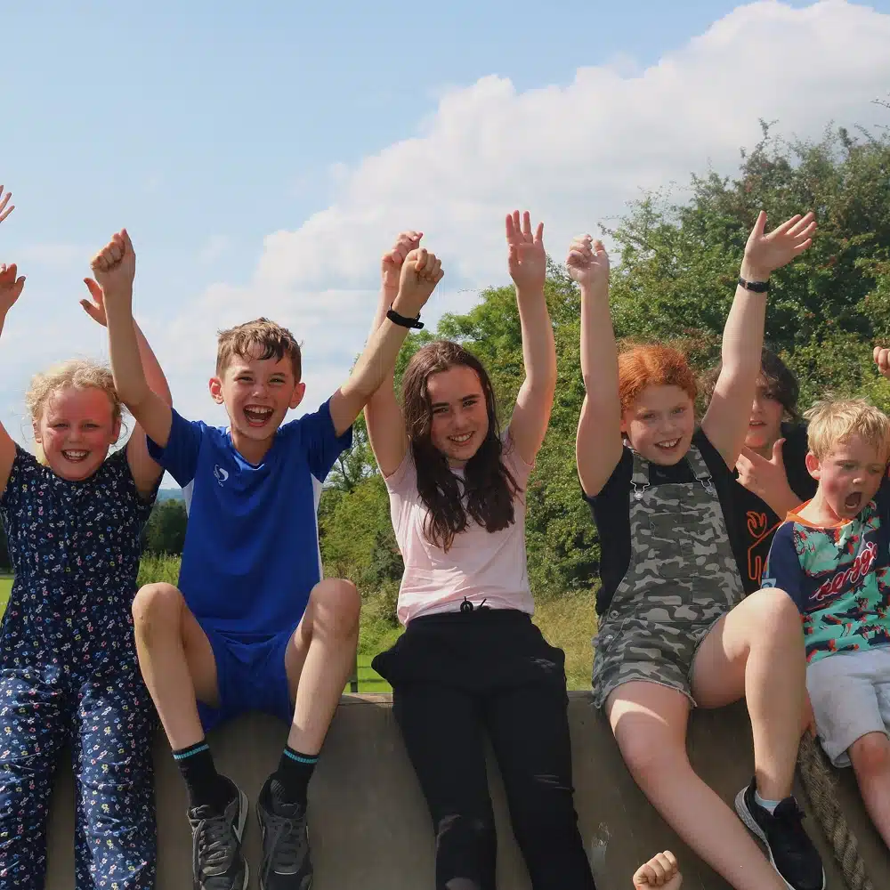 Six children sit on a low wall outdoors, smiling and raising their arms joyfully. Trees and blue sky are in the background, and the children appear to be having fun together on a sunny day.