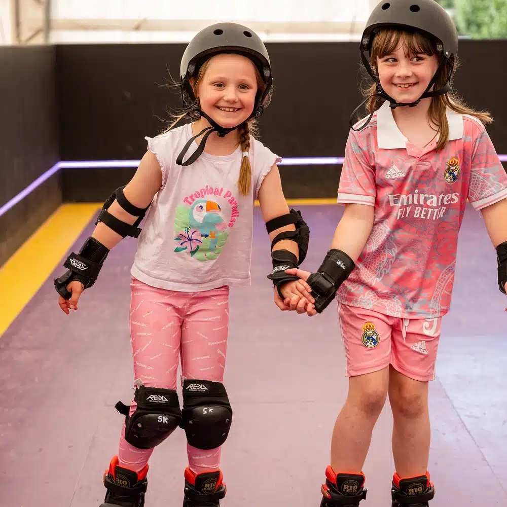 Two young girls wearing helmets, knee pads, and roller skates hold hands and smile while skating indoors. One wears a tropical bird shirt and pink leggings; the other wears a pink football jersey and shorts.
