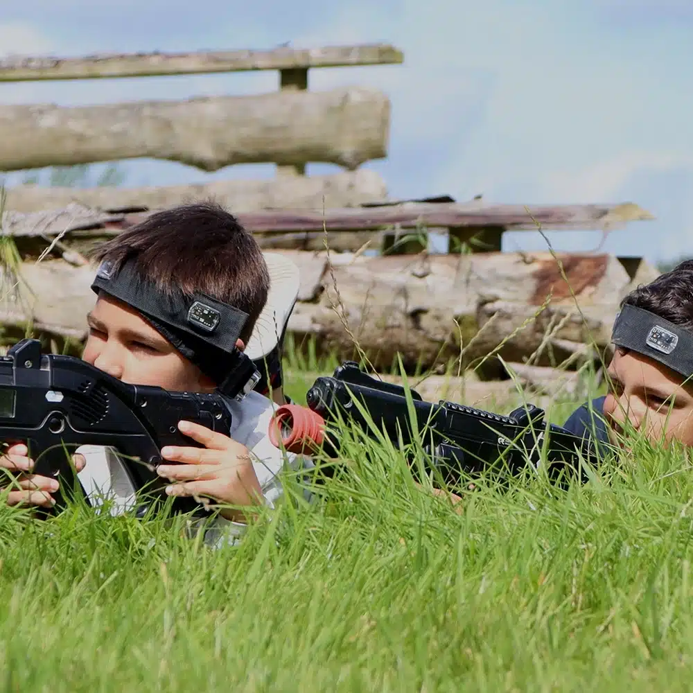 Two children wearing headbands and holding laser tag guns lie in tall grass outdoors, aiming ahead. Wooden logs and a clear sky are visible in the background.
