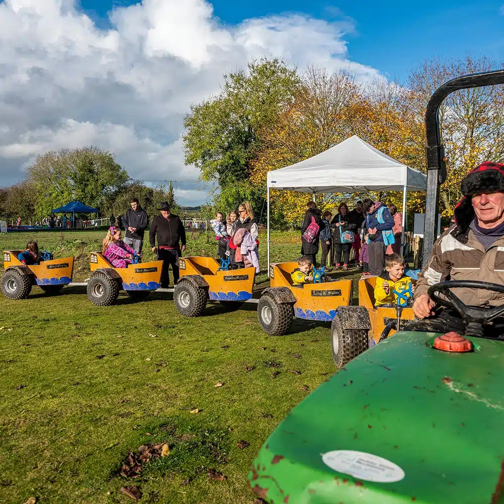 Children ride in small, yellow, tractor-pulled carts while adults supervise nearby. Other people gather under a white marquee. The scene is set outdoors on grass with trees and a partly cloudy sky.