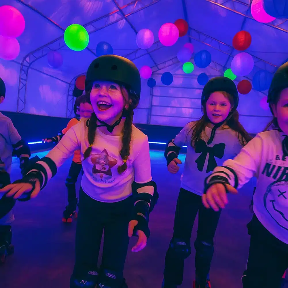 Children wearing helmets and roller skates smile and skate indoors under colourful hanging lights and lanterns, creating a joyful and playful atmosphere.