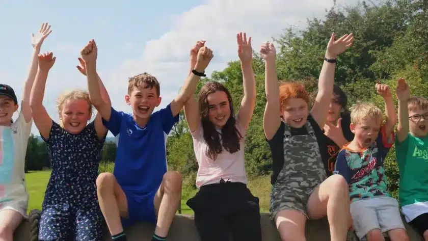 Seven children sit on a low wall outdoors at Navan summer camp, smiling and raising their arms in celebration. They appear happy and joyful, with green trees and a blue sky in the background.