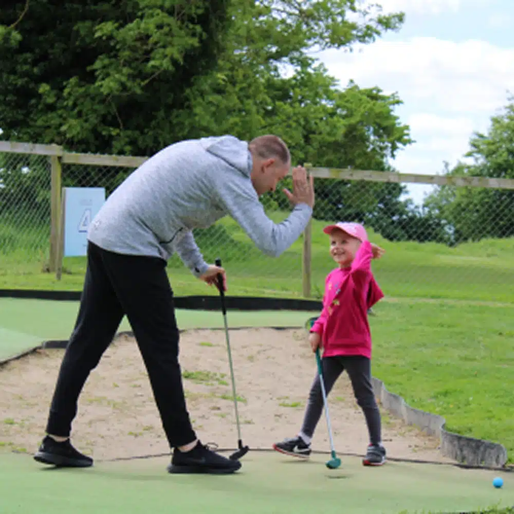 A man and a young girl enjoy crazy golf outdoors, sharing a high five during their Navan family activities. The man, golf club in hand, bends to her level as she smiles in a pink jacket and hat. Green grass and trees complete the scene.