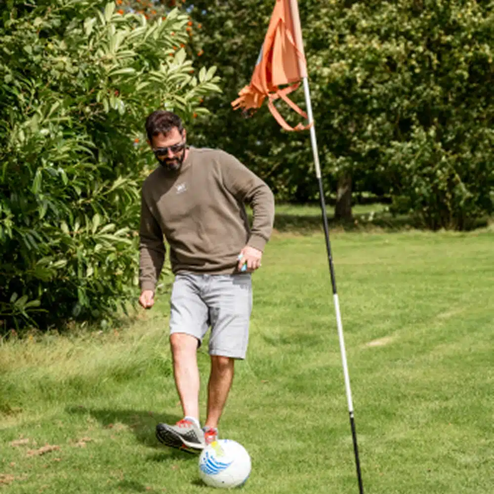 A man wearing sunglasses, a green sweatshirt, and grey shorts kicks a football near a flag on a grassy field, surrounded by trees—perfect for those enjoying Navan family activities.