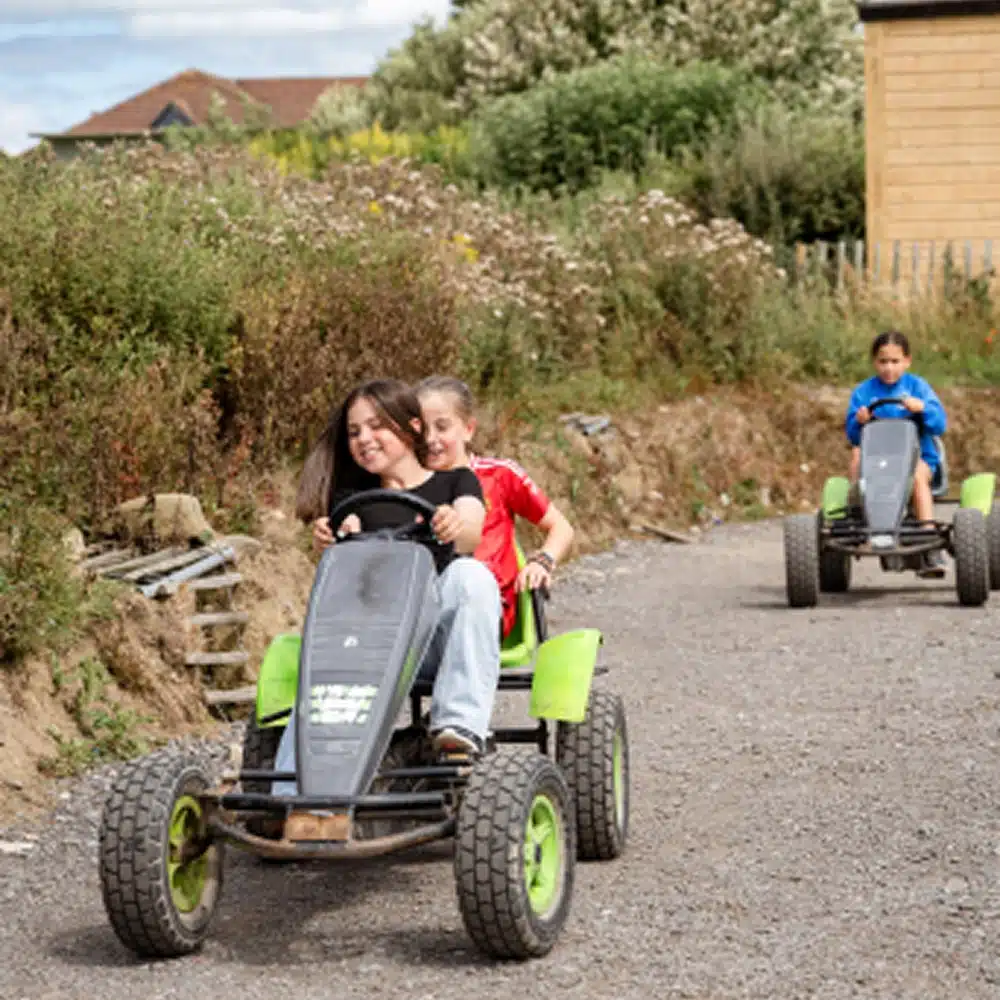 Two children ride together in a pedal go-kart on a gravel path, smiling and having fun—a perfect example of Navan family activities—whilst another child follows behind. Bushes, trees, and a wooden building can be seen in the background.