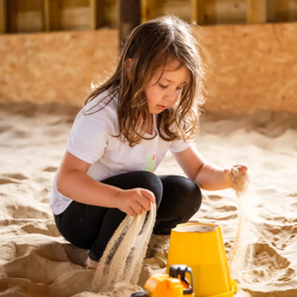A young child with long brown hair sits in a sandpit indoors, letting sand fall through her hands next to a yellow bucket and toy lorry—enjoying one of many fun Navan family activities. She is wearing a white shirt and black trousers.