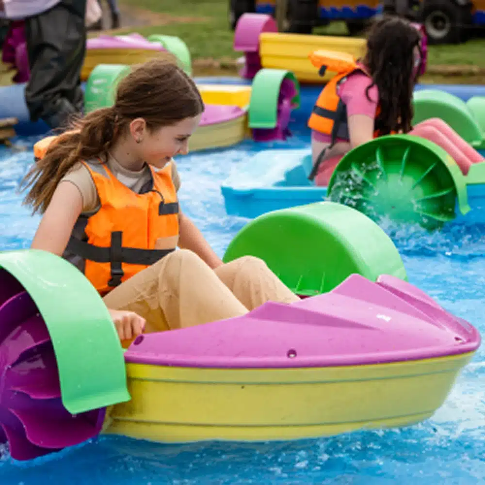 Two children wearing orange life jackets ride colourful pedal boats in a small pool, enjoying one of the many Navan family activities. The water is blue, and the bright yellow, purple, and green boats add to their excitement and smiles.