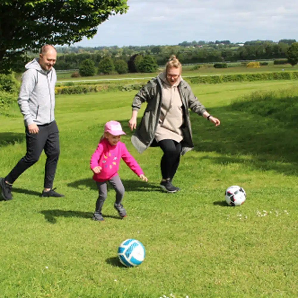 A man, woman, and young child are enjoying Navan family activities as they play football together on a grassy field under a tree, with two footballs visible and fields in the background.