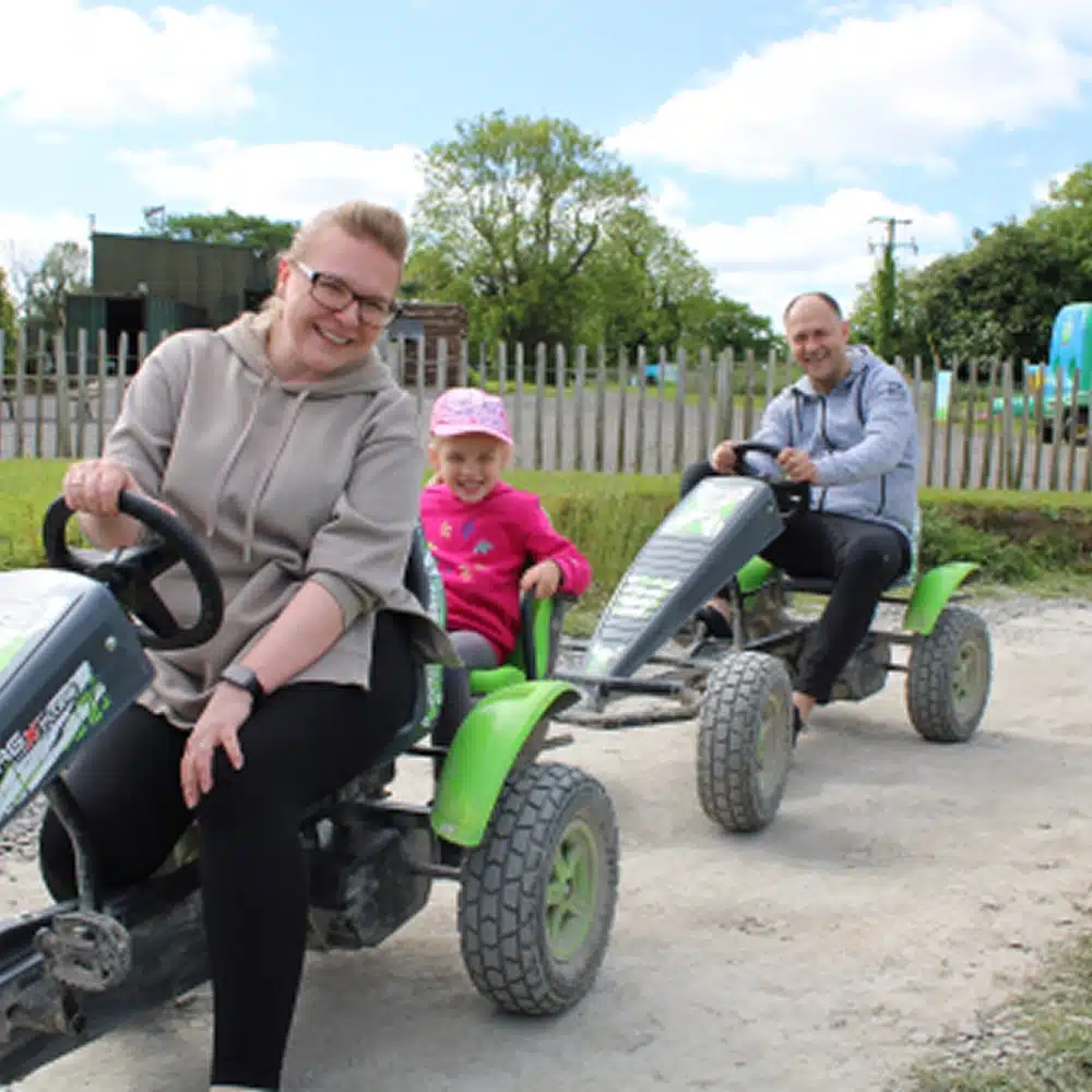 A woman, a young girl, and a man are smiling and riding green pedal go-karts on a dirt path outdoors—enjoying one of the fun Navan family activities, with a wooden fence and trees in the background.