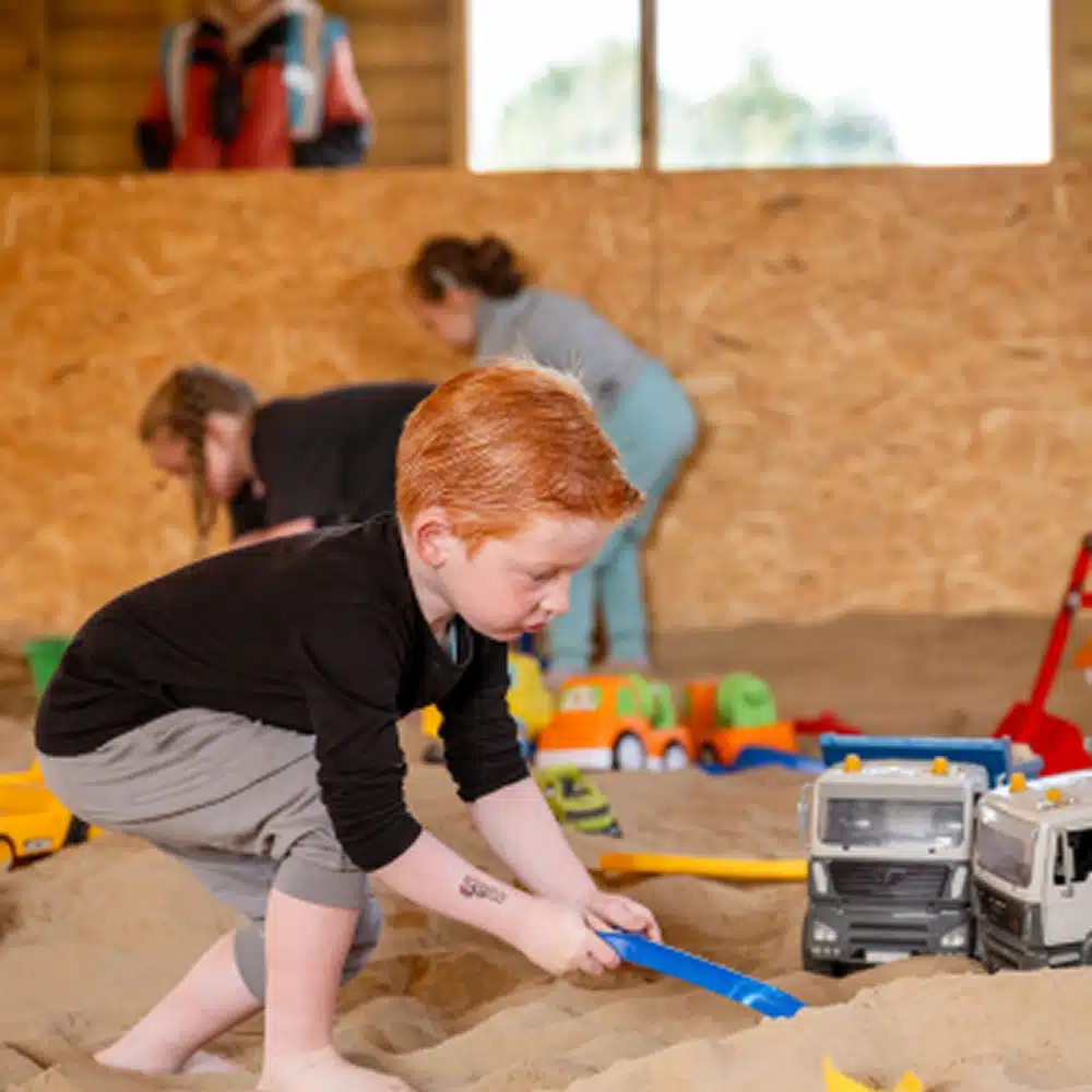 A young boy with red hair enjoys Navan family activities, playing in an indoor sandpit and digging with a blue spade near toy lorries, while other children play and interact in the background.