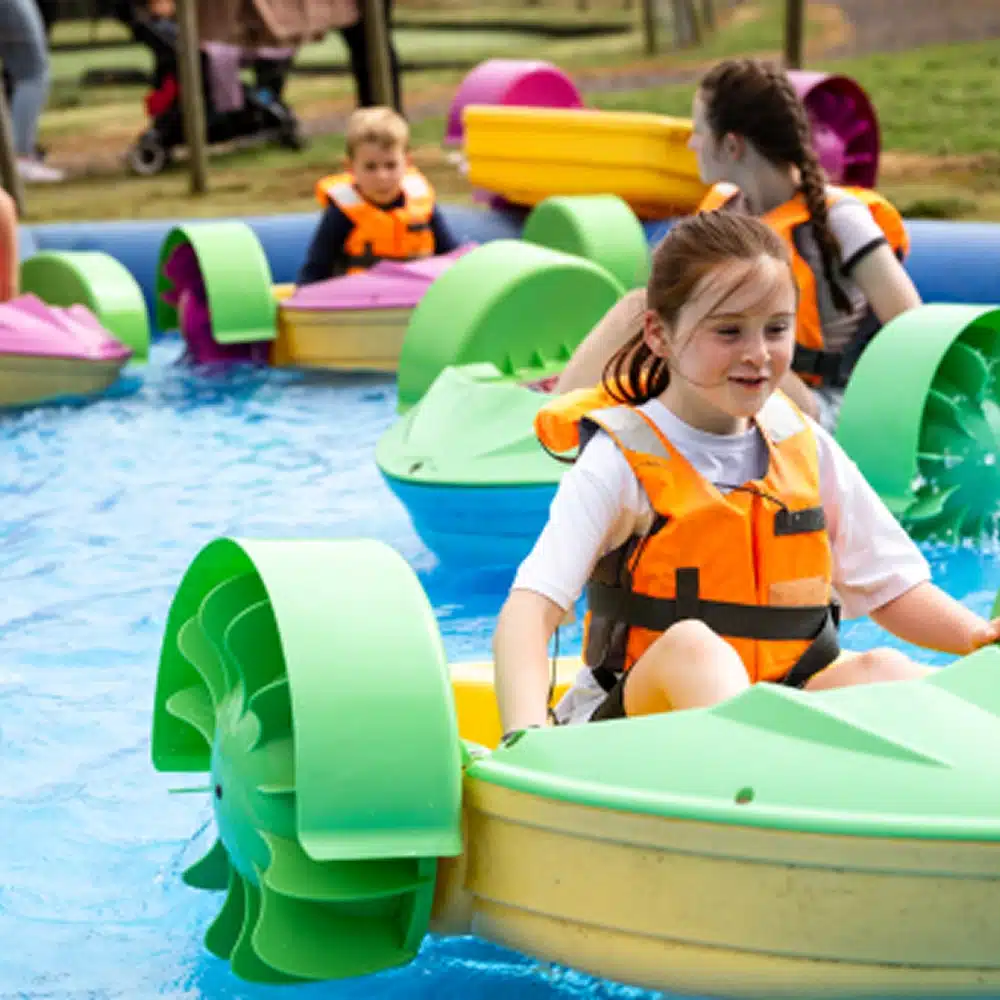 Children wearing life jackets paddle small colourful boats in a shallow pool, enjoying a sunny day outdoors—one of the fun Navan family activities. Other children and adults are visible in the background near the edge of the pool.