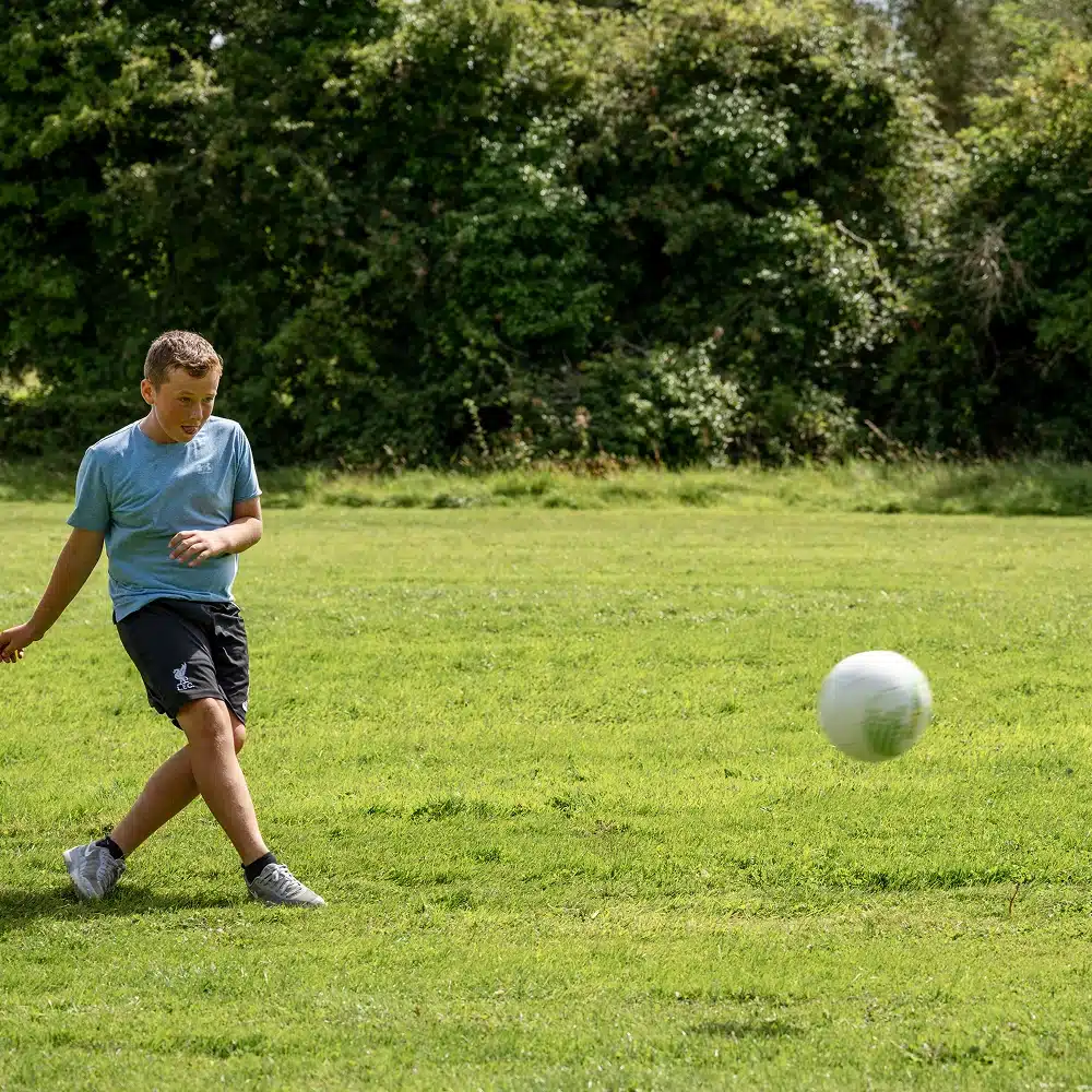 A boy in a blue shirt and black shorts kicks a white football on a grassy field with trees in the background, enjoying one of the many Navan winter family activities on a sunny day.