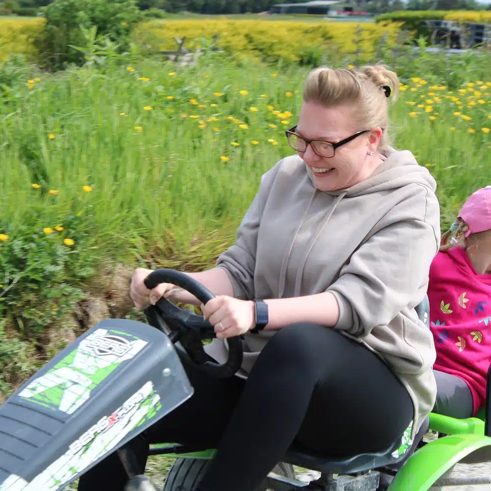 A woman wearing glasses and a grey hoodie smiles whilst riding a green pedal go-kart outdoors with a child in a pink cap. Yellow wildflowers and grass fill the background, capturing the joy of Navan winter family activities.