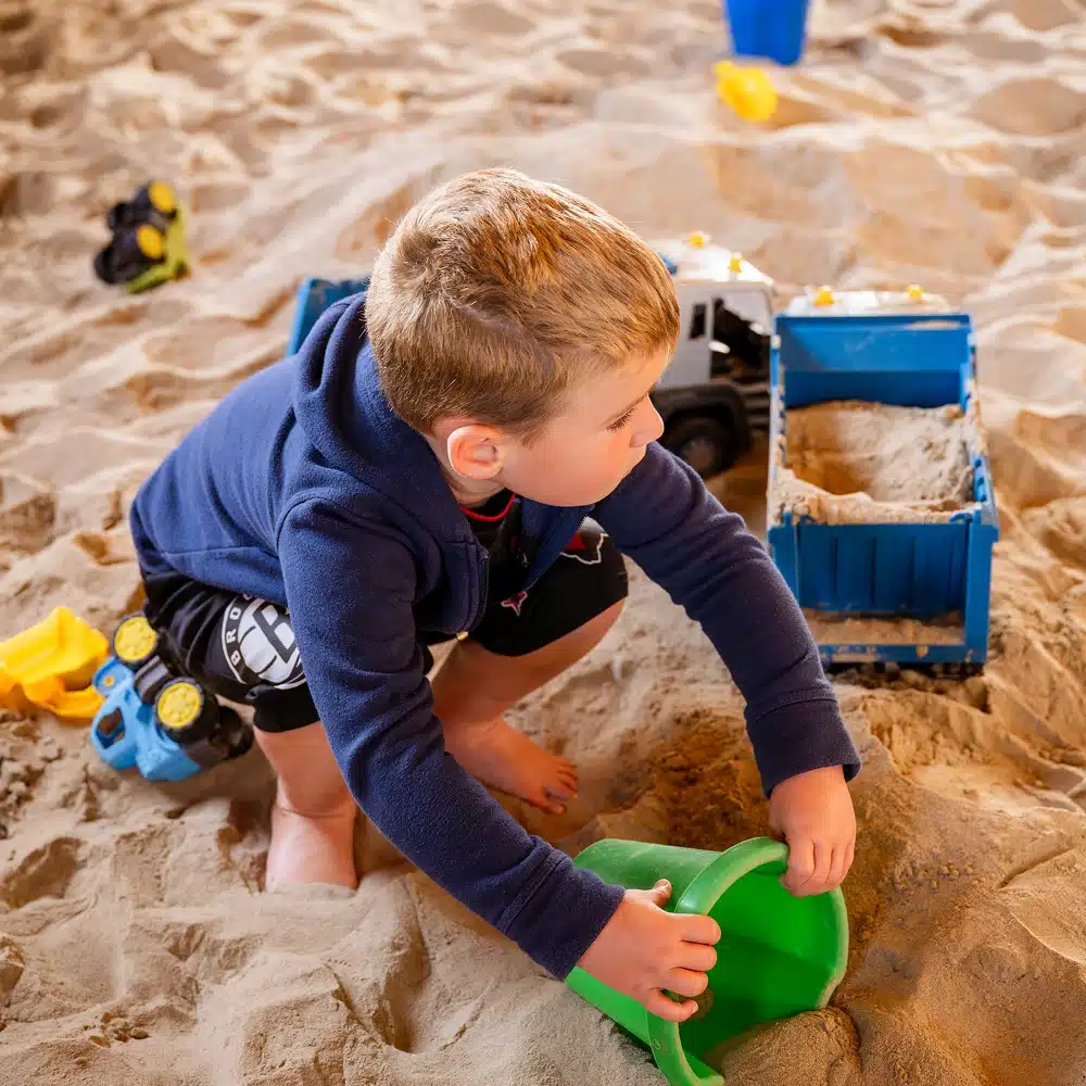 A young boy wearing a blue jacket plays in a sandpit, scooping sand into a green bucket. Toy lorries and a blue sand container are nearby. The scene captures the joy of Navan winter family activities, with the boy barefoot and focused on his play.