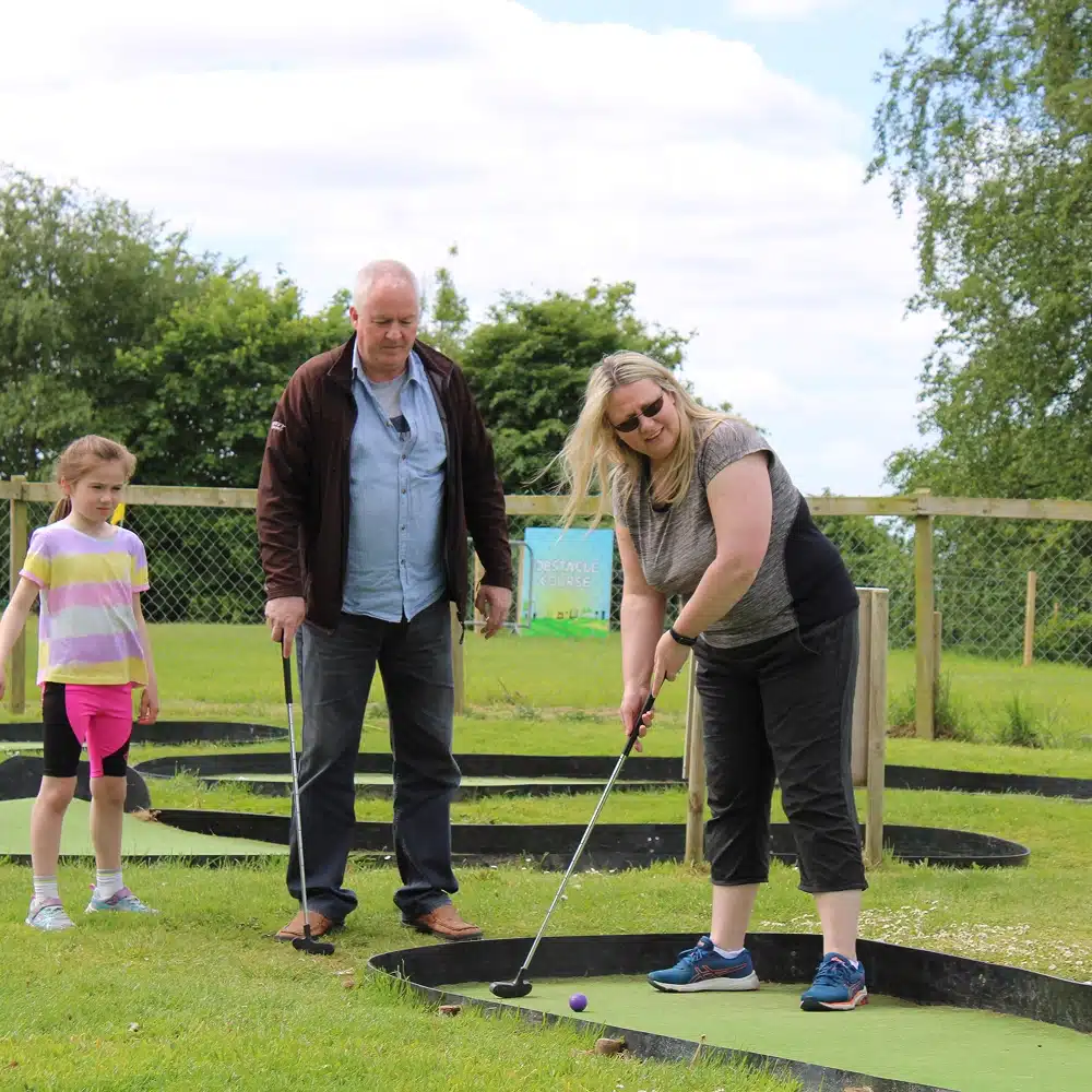 A smiling woman plays crazy golf as a man and a young girl watch. They are outdoors on a grassy course in Navan, enjoying winter family activities under a partly cloudy sky with trees and a fence in the background.