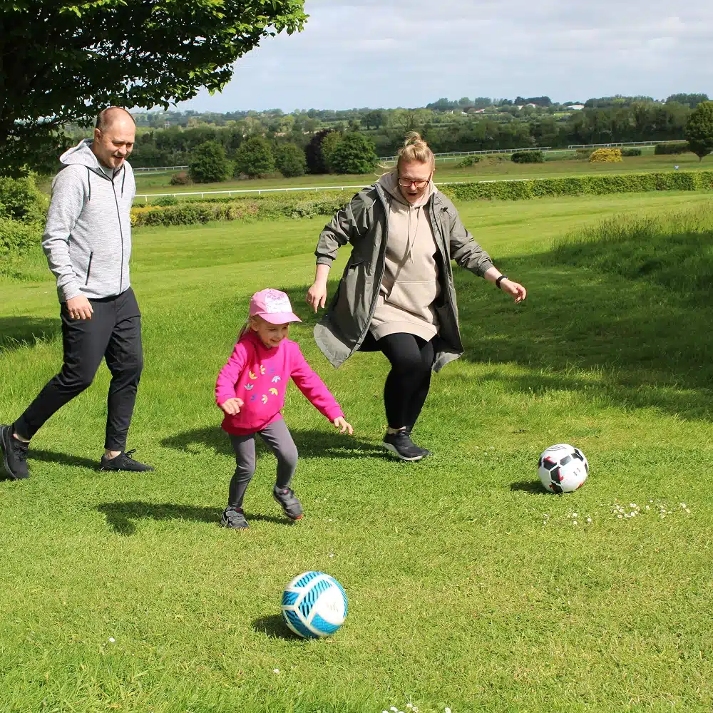A man, woman, and young girl play football together on a grassy field—a fun example of Navan winter family activities. The girl, in a pink jacket and cap, runs near the ball as the adults follow, smiling under a cloudy sky with trees in the background.