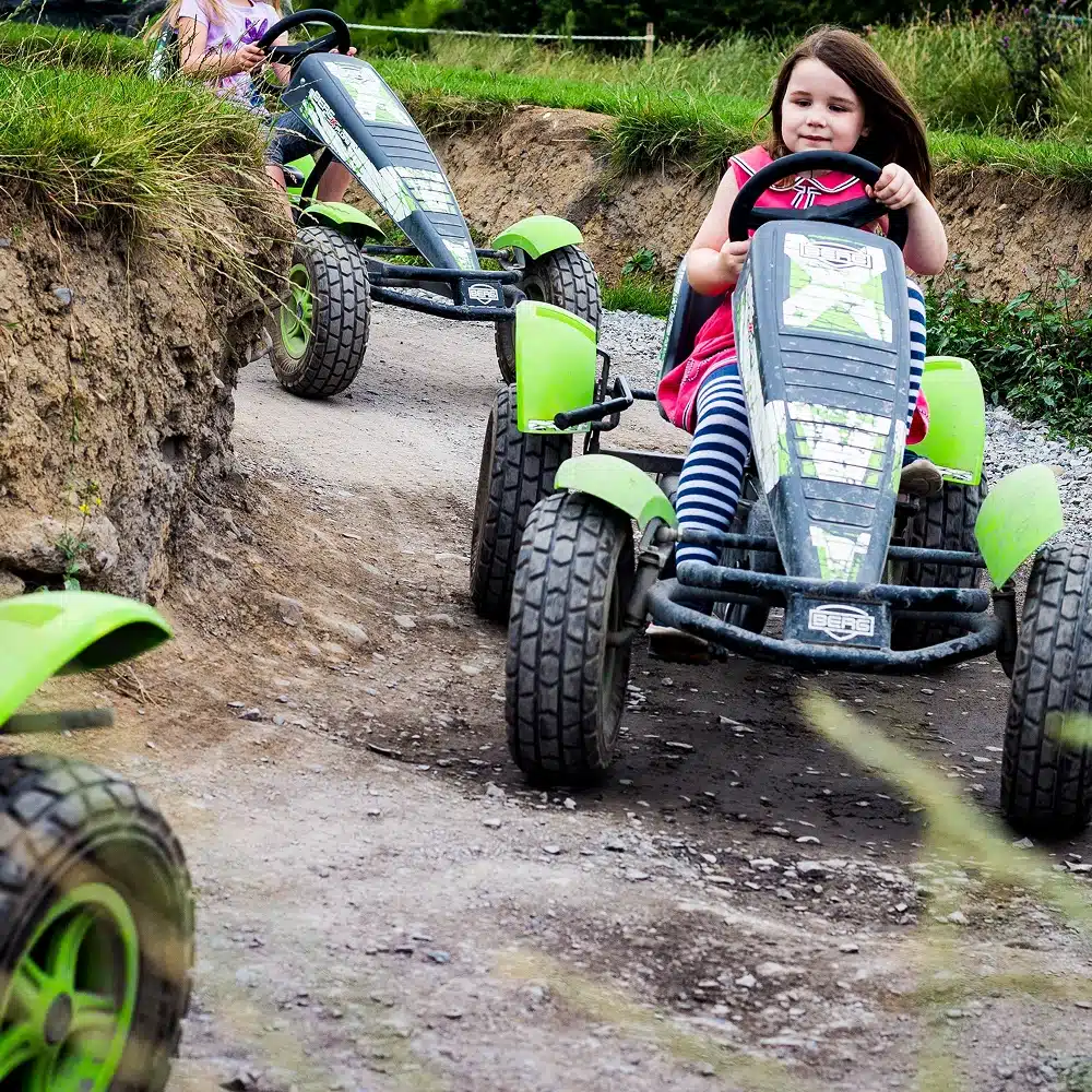 A young girl with brown hair drives a green pedal go-kart on a dirt track, smiling in her pink hoodie. Another child follows close behind. This fun outdoor scene is perfect for Navan winter family activities among the grassy banks.