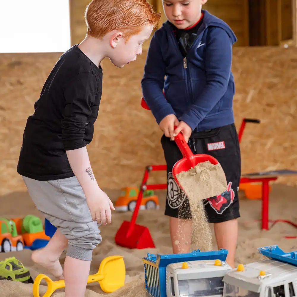 Two young boys enjoy Navan winter family activities, playing in a sandpit with toy lorries and spades. One pours sand from a red spade into a toy tipper lorry as the other watches, with colourful vehicles scattered in the background.