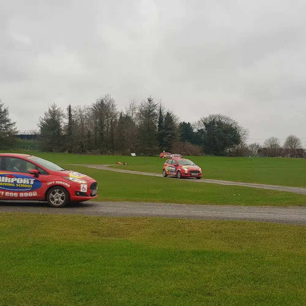 Two red cars with driving school logos drive on a narrow road through a grassy park area, with trees and cloudy sky in the background—an ideal scene for Navan youth group activities centred around safe driving.