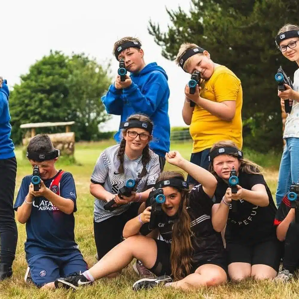 A group of children outdoors, wearing headbands and holding laser tag guns, pose together and smile at the camera on a grassy field—enjoying Navan youth group activities with trees and outdoor structures in the background.