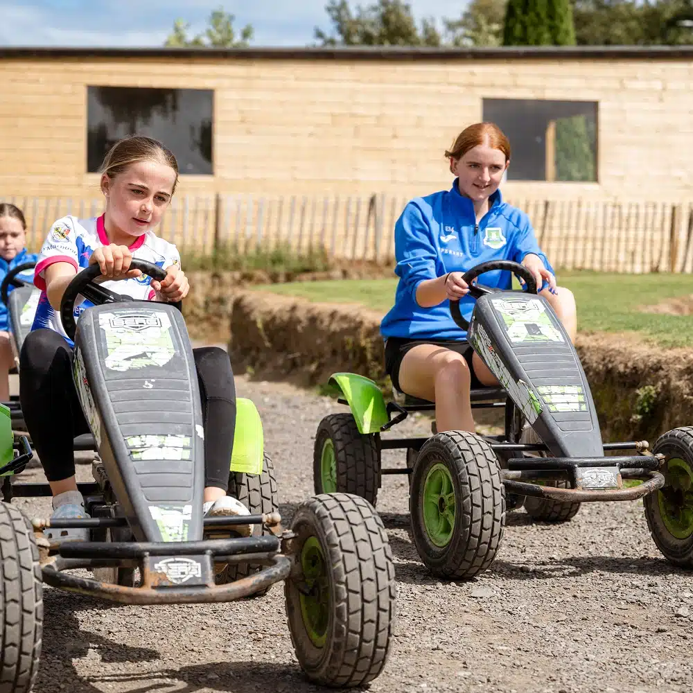 Two girls are riding pedal go-karts on a gravel track outdoors, both focused on steering. Other children and a wooden building are visible in the background—one of many fun Navan youth group activities on this sunny day.