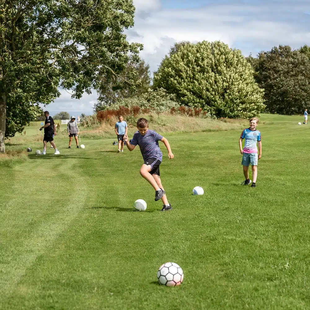 Several boys, part of Navan youth group activities, play football on a grassy field surrounded by trees. One boy in front is about to kick a football while others stand or walk in the background with more balls.