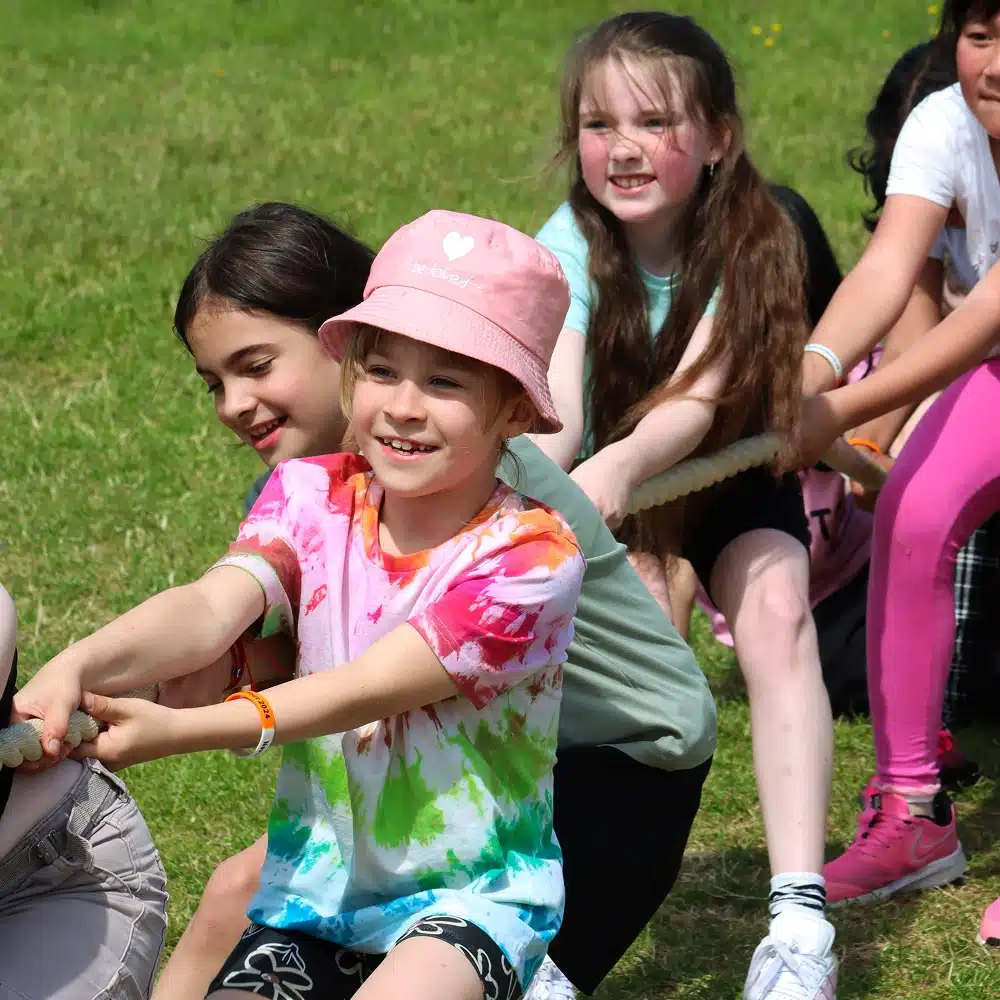 A group of smiling children outdoors, pulling together on a rope in a tug-of-war game—part of Navan youth group activities. The child at the front wears a pink hat and tie-dye shirt, with green grass suggesting a sunny, playful day.