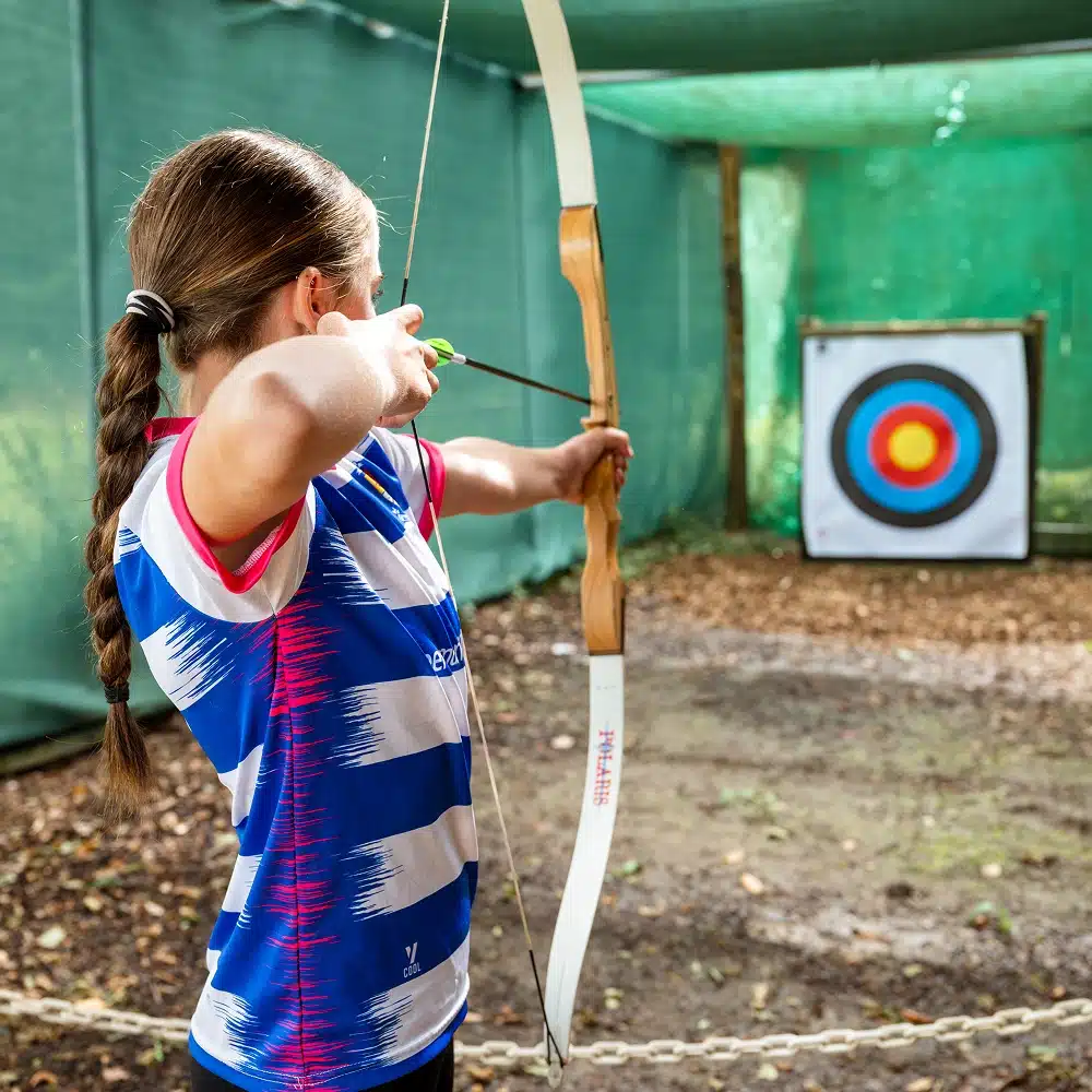 A girl with a plait aims a bow and arrow at a colourful target in an outdoor archery range, wearing a blue and white striped shirt—part of Navan Secondary School's laser tag and activity day.