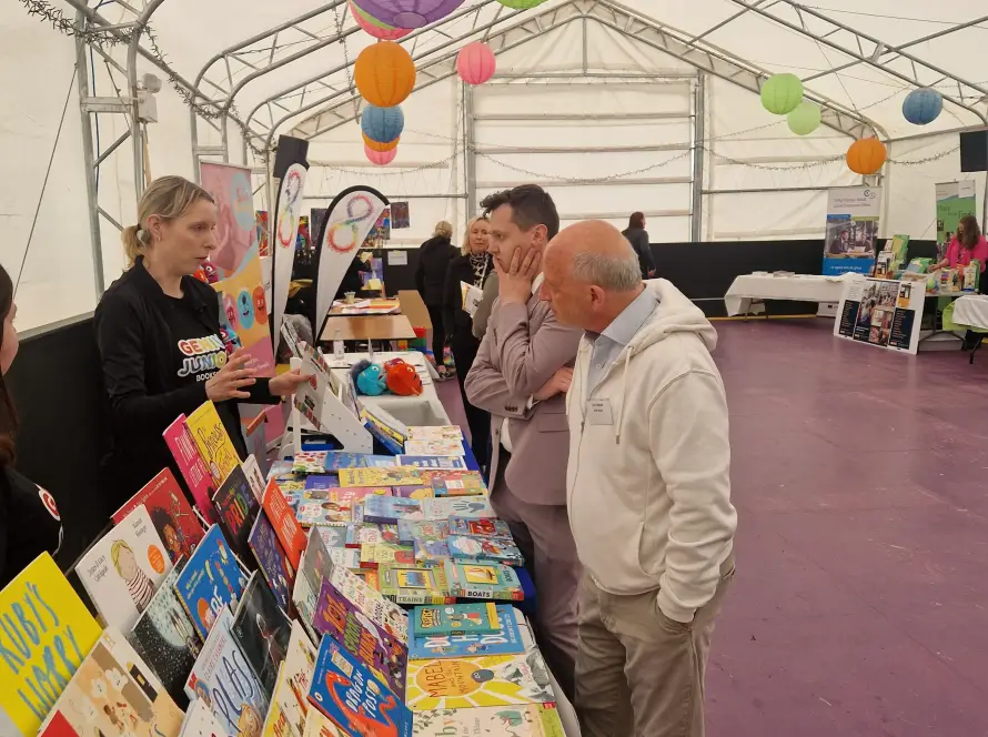 A woman stands behind a table displaying colourful children's books, talking to two men in a large white marquee decorated with hanging lanterns and filled with various stalls.