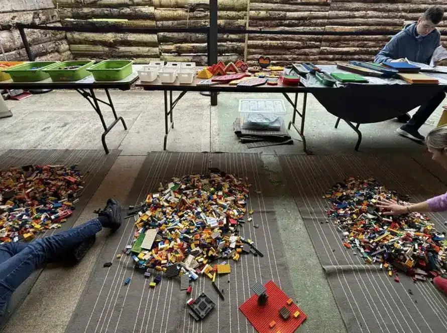 A room with three rugs covered in scattered LEGO bricks, two people lying or sitting on the mats building, and tables in the background holding sorted LEGO pieces and trays. Colourful fabric panels hang above a wooden fence.