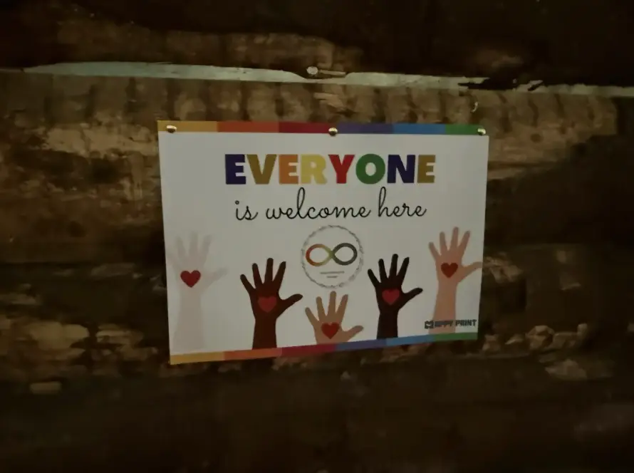 A sign on a wooden wall reads, "EVERYONE is welcome here," with colourful letters and illustrations of diverse raised hands with hearts on the palms. There is also an infinity symbol in the centre.