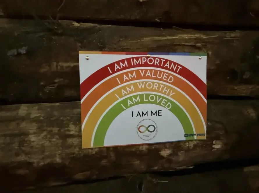 A colourful sign on a wooden wall displays rainbow arcs with the words: "I AM IMPORTANT, I AM VALUED, I AM WORTHY, I AM LOVED, I AM ME." An infinity symbol appears at the bottom.