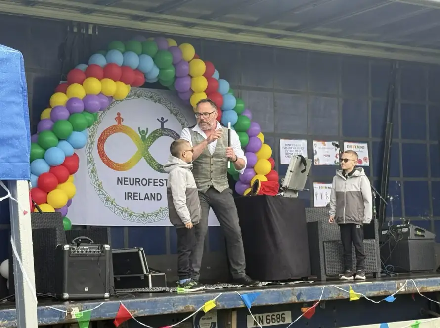 A magician performs a trick with two young boys on stage at Neurofest Ireland. The stage is decorated with colourful balloons, a festival banner, and various equipment, whilst the boys watch the magician intently.