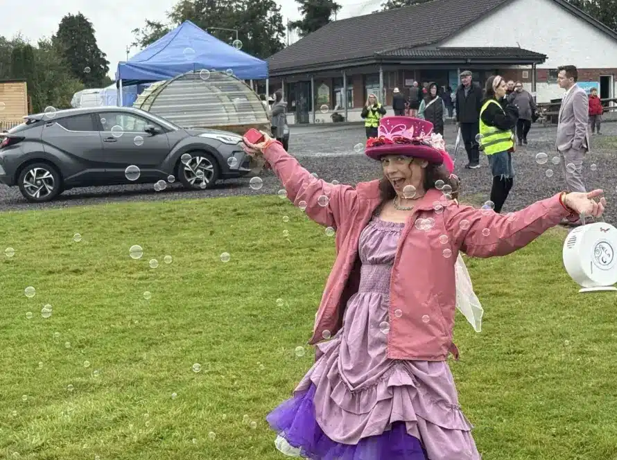 A woman in a pink hat and purple dress joyfully spreads her arms on a grassy field, surrounded by bubbles. People and cars are in the background near a building and blue marquee on a cloudy day.