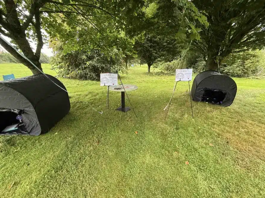 Two black tents are set up on grass under large trees, with a small round table and two signs between them. The surrounding area is green and lush with trees in the background.